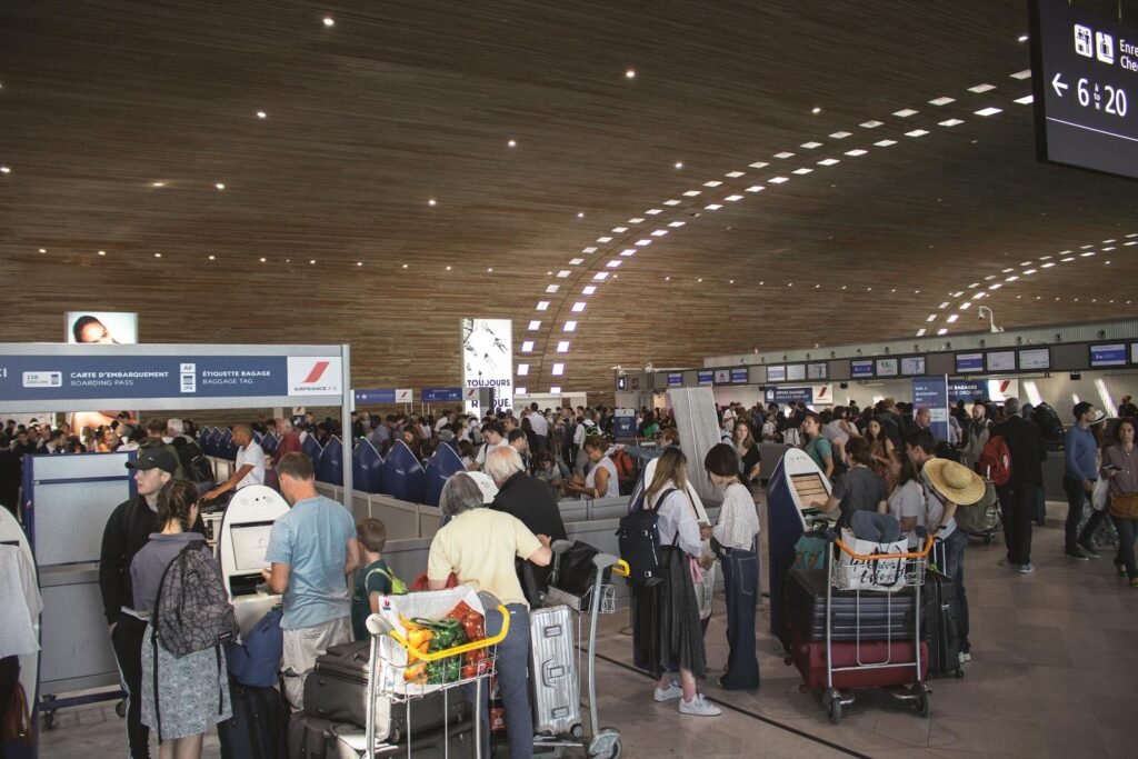A stunning, ultra-realistic photograph of a busy airport terminal, with travelers rushing through the vast, airy space. The foreground features a large departures board, displaying a variety of flight options and destinations, including several to major European cities. Sleek, modern architectural elements, such as floor-to-ceiling windows and metallic accents, create a sense of efficiency and modernity. In the middle ground, groups of passengers can be seen navigating their way through security checkpoints and boarding gates, their expressions a mix of excitement and anticipation for their European adventures. The background is filled with the activity of an international airport, with airport staff, luggage carts, and the occasional glimpse of an aircraft taking off or landing, all bathed in warm, natural lighting that filters in through the expansive windows. Crowded airport terminal in europe with travellers in line. Indoor setting with modern architecture.