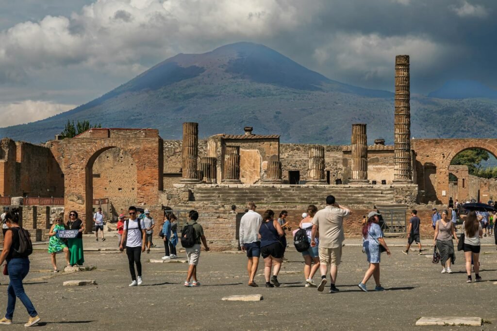 A bustling entrance to the ancient ruins of Pompeii, with visitors queuing at the ticket booths and navigating the pathways leading into the archaeological site. The foreground depicts the flow of people disembarking from buses and taxis, their luggage in hand, while the middle ground showcases the grand, weathered entrance gates. In the background, glimpses of the well-preserved ruins and the iconic Mount Vesuvius loom, creating a sense of anticipation and wonder. Warm Mediterranean light bathes the scene, adding depth and a palpable sense of history. An ultra-realistic photo capturing the logistical and atmospheric experience of embarking on a Pompeii day tour from Naples or Rome. A bustling entrance to the ancient ruins of Pompeii, with visitors queuing at the ticket booths and navigating the pathways leading into the archaeological site. The foreground depicts the flow of people disembarking from buses and taxis, their luggage in hand, while the middle ground showcases the grand, weathered entrance gates. In the background, glimpses of the well-preserved ruins and the iconic Mount Vesuvius loom, creating a sense of anticipation and wonder. Warm Mediterranean light bathes the scene, adding depth and a palpable sense of history. An ultra-realistic photo capturing the logistical and atmospheric experience of embarking on a Pompeii day tour from Naples or Rome.