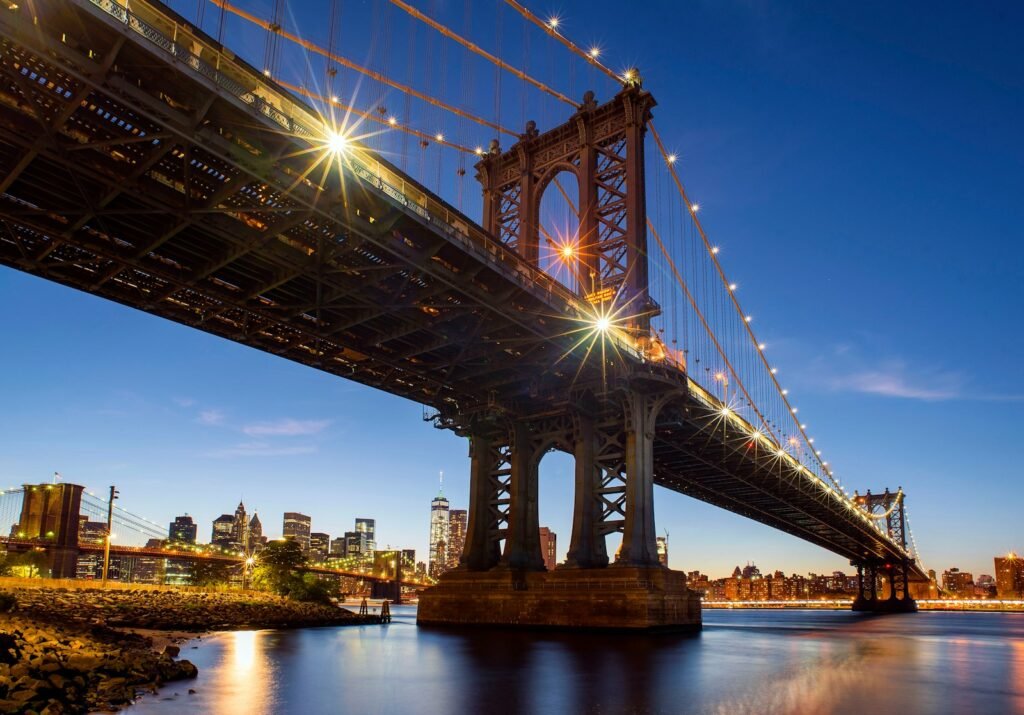 Iconic skyline of Lower Manhattan, with the towering silhouettes of the Freedom Tower and other skyscrapers, reflected in the calm waters of the East River. In the foreground, the elegant suspension bridge of the 1 Hotel Brooklyn Bridge, its modern architecture seamlessly blending with the historic cityscape. The scene is bathed in warm, golden light, casting a serene and inviting atmosphere. The image is captured from a vantage point on the Brooklyn waterfront, showcasing the breathtaking panorama of New York's most iconic landmarks. Iconic skyline of Lower Manhattan, with the towering silhouettes of the Freedom Tower and other skyscrapers, reflected in the calm waters of the East River. In the foreground, the elegant suspension bridge of the 1 Hotel Brooklyn Bridge, its modern architecture seamlessly blending with the historic cityscape. The scene is bathed in warm, golden light, casting a serene and inviting atmosphere. The image is captured from a vantage point on the Brooklyn waterfront, showcasing the breathtaking panorama of New York's most iconic landmarks.
