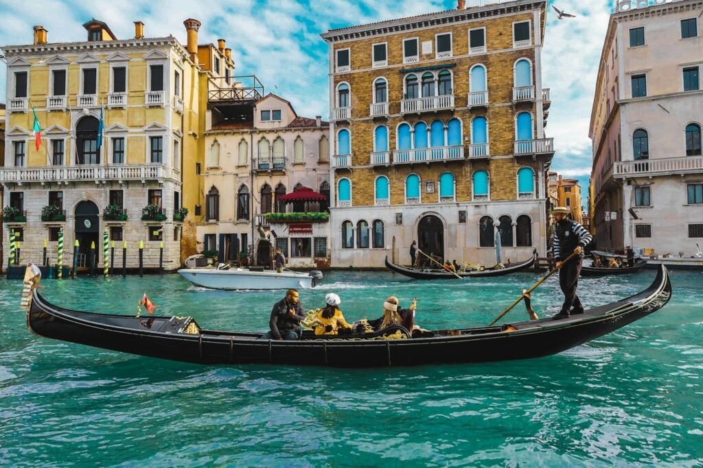 A dreamlike dawn illuminates the serene canals of Venice, Italy. Elegant gondolas glide effortlessly through the still waters, their carved wooden hulls reflecting the soft, golden light. Centuries-old architecture lines the canals, their ornate facades mirrored in the tranquil surface. Wispy clouds drift overhead, casting soft shadows that dance across the scene. The atmosphere is one of tranquility and timeless beauty, inviting the viewer to experience the magic and wonder of this iconic European destination. people on boat in Venice Italy, also one of the best cities in europe to visit