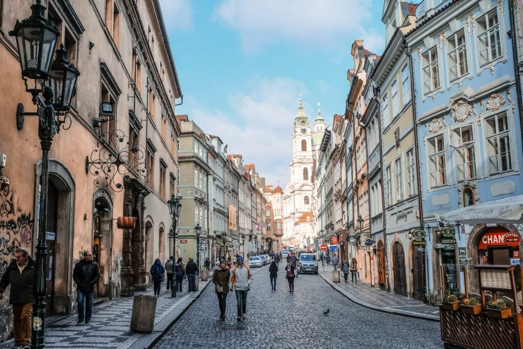 A picturesque European city nestled in rolling hills, with charming cobblestone streets and colorful buildings. The foreground features a winding road leading into the quaint town center, with locals going about their daily routines. In the middle ground, a sprawling medieval cathedral stands tall, its gothic spires piercing the sky. In the background, verdant forests and mountains create a serene, untouched backdrop. Warm, natural lighting bathes the scene, highlighting the timeless beauty of this off-the-beaten-path destination. An ultra-realistic, cinematic photo capturing the essence of budget-friendly European travel. European city with colorful buildings. The foreground features a road leading into the quaint town center, with locals going about their daily routines. The photo capturing the essence of budget-friendly European travel.