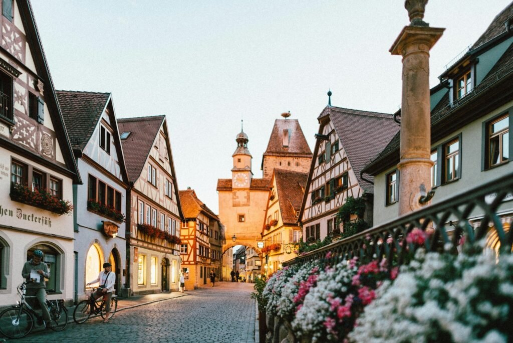 A bustling street in a quaint European city, lined with charming cafes and local boutiques. The sun casts a warm, golden glow, highlighting the rich, earthy tones of the buildings. In the foreground, a group of travelers sip coffee and savor the local cuisine, enjoying the off-season tranquility. In the middle ground, a couple strolls hand-in-hand, admiring the historic architecture. In the background, a picturesque town square comes into view, showcasing the shoulder-season beauty of this affordable European destination. white and pink petaled flowers on metal fence near concrete houses and tower at daytime in europe