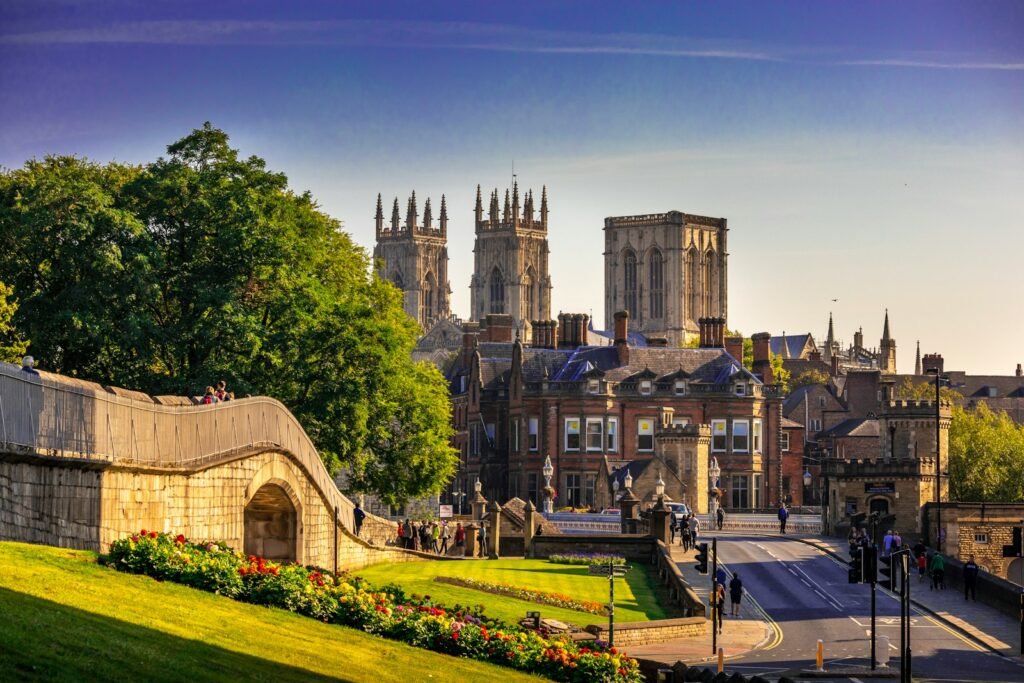 A sun-dappled medieval city skyline, York's ancient walls and iconic Minster cathedral loom majestically. In the foreground, the charming timber-framed buildings of The Shambles, a historic cobblestone market street, invite exploration. Warm natural lighting casts a golden glow, highlighting the intricate architectural details. The scene evokes a sense of timeless elegance and tranquility, as if frozen in a moment from centuries past. Shot with a wide-angle lens to capture the full grandeur, this ultra-realistic photo showcases the captivating blend of York's medieval heritage and enduring splendor. York, England, brown and white concrete building near green trees during daytime