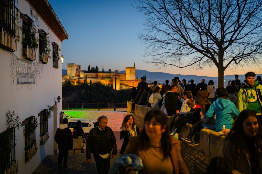 A grandiose vista of Granada's Alhambra palace, its intricate Moorish arches and dazzling courtyards basking in warm, golden light. In the distance, the soaring spires of Seville's Alcázar castle pierce the azure sky, framed by lush, verdant gardens. In the foreground, a flamenco dancer twirls passionately, her vibrant crimson dress flowing with the rhythm of the music, capturing the captivating essence of Andalusia's cultural soul. The scene is imbued with a sense of timeless elegance and historical grandeur, inviting the viewer to immerse themselves in the captivating beauty of these iconic Iberian destinations. a group of people walking down a street next to a building in Granada Spain