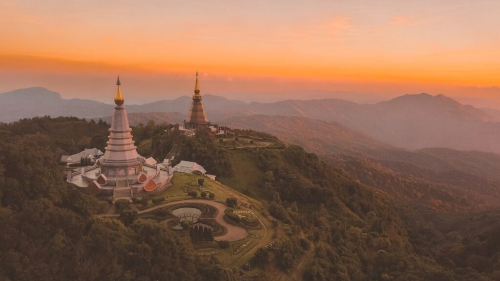 A serene mountainous landscape in northern Thailand, bathed in warm golden light. In the foreground, a winding road cuts through terraced rice paddies, with farmers tending to the lush green fields. In the middle ground, traditional wooden houses and ornate Buddhist temples dot the rolling hills, their roofs glinting in the sunlight. In the distance, majestic peaks rise up, their jagged silhouettes framed by a vibrant, painted sky. The scene exudes a sense of tranquility and cultural richness, inviting the viewer to explore the depth and diversity of Thailand beyond its famous beaches.