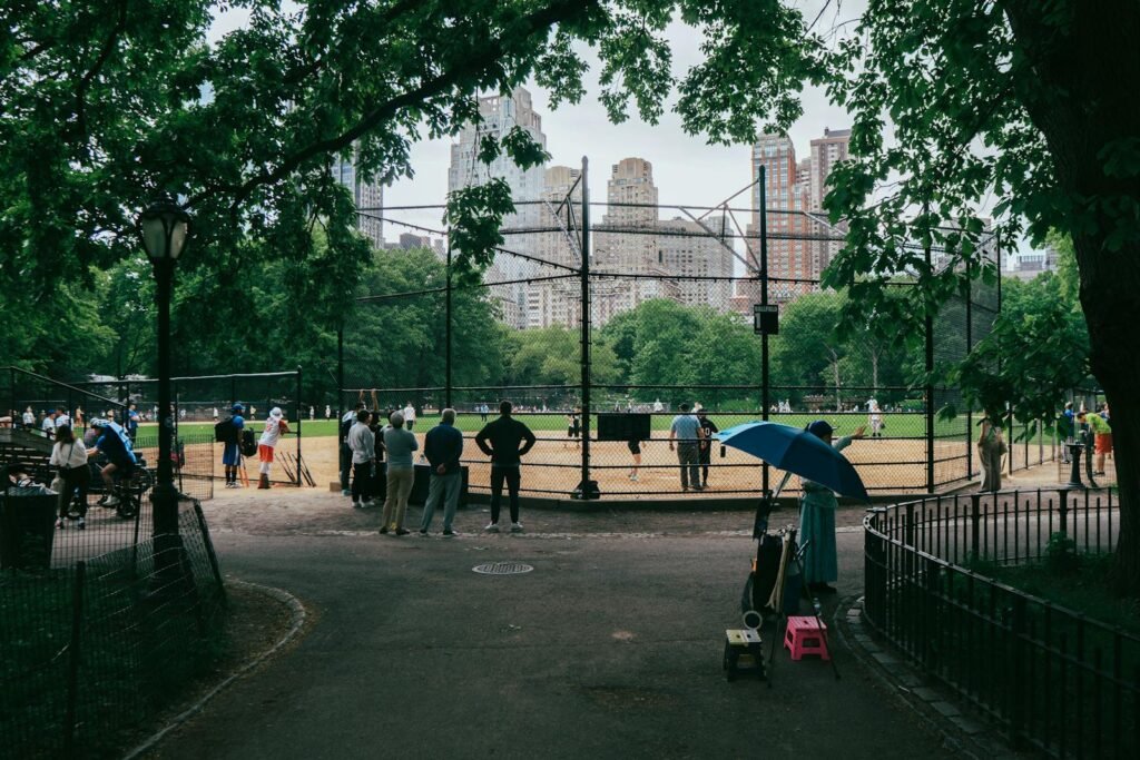 A bustling city skyline with iconic landmarks like the Empire State Building and the Statue of Liberty in the background, framed by a vibrant, kid-friendly foreground. In the middle ground, families explore diverse attractions like the Central Park Zoo, the American Museum of Natural History, and the Intrepid Sea, Air & Space Museum. Colorful playground equipment, ice cream vendors, and street performers create a lively, energetic atmosphere. The scene is bathed in warm, golden sunlight, conveying a sense of wonder and adventure for children and their parents to discover the endless possibilities of New York City. A baseball game in Central Park with Manhattan skyscrapers in the background. 