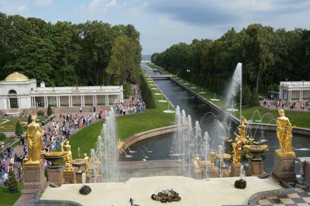 A view of a park with fountains and people panorama of historic European landscapes, bathed in warm, golden sunlight. In the foreground, a group of travelers stand in awe, captivated by the imposing ruins of a grand castle, its weathered stone walls and towers a testament to the region's storied past. In the middle ground, a winding road leads through picturesque countryside, dotted with quaint villages and rolling hills. In the distance, the silhouettes of soaring mountains rise against a brilliant blue sky, hinting at the vast, untamed beauty that awaits. Cinematic camera angles, with a focus on depth and perspective, invite the viewer to step into this evocative scene, to experience the allure of exploring Europe's enduring World War II heritage firsthand.