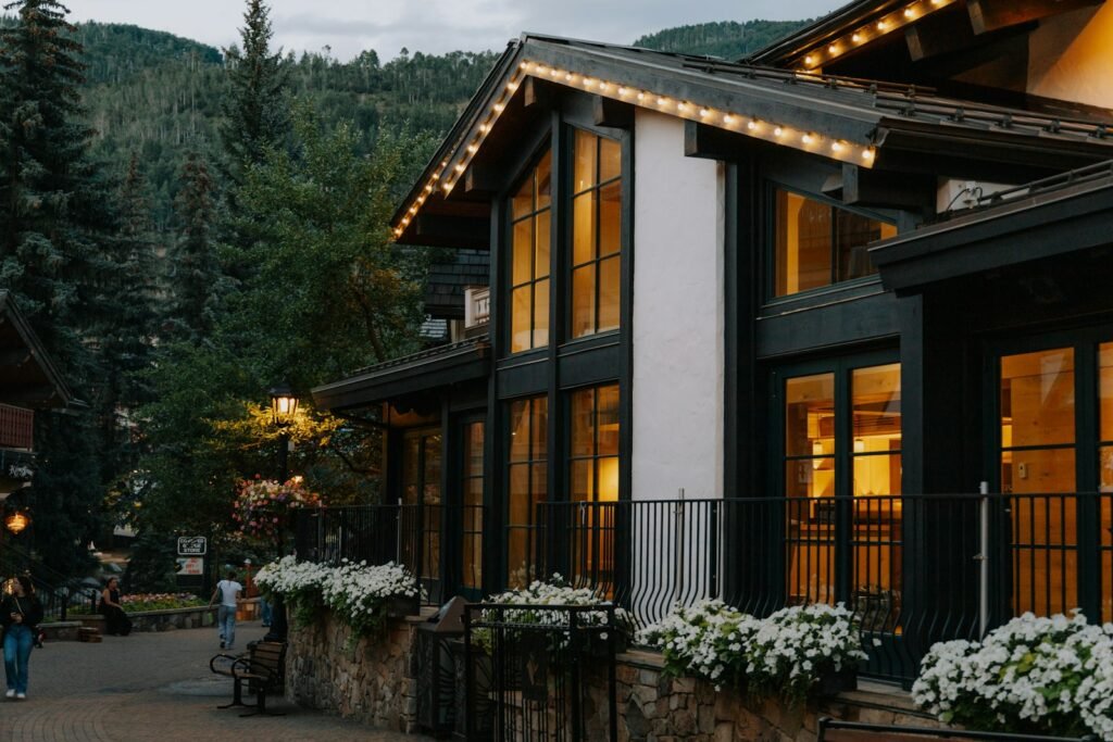 Serene Catskill oasis: a panoramic shot of an elegant, multi-story hotel nestled amid lush, verdant hills. Its sleek, modern facade gleams in the warm, golden afternoon light, while a sparkling, azure pool occupies the foreground, framed by well-manicured gardens and swaying trees. Guests can be seen lounging poolside, soaking up the tranquil atmosphere. The scene exudes a sense of relaxation and rejuvenation, perfectly capturing the essence of a luxurious Catskill Mountain getaway.