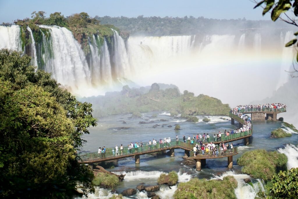 A vibrant photo showcasing the allure of South America tours from the USA. In the foreground, a group of adventurous travelers walking on the bridge and gaze in awe at the majestic Iguazu Falls, its thunderous cascades framed by lush, verdant foliage. In the middle ground, a bustling city skyline rises, reflecting the diverse cultures and architecture of the region. The background features snow-capped Andean peaks, hinting at the breathtaking landscapes and outdoor activities that await. Warm, golden lighting bathes the scene, evoking the warmth and excitement of a South American vacation. Captured through a wide-angle lens, the image conveys the grandeur and scale of the ultimate South America tour experience.