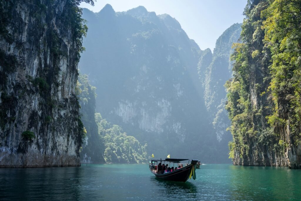 An idyllic riverside scene in Laos, with a traditional wooden boat drifting down a winding jungle-fringed river. In the foreground, a pair of locals relax on the boat's deck, taking in the lush, verdant landscape. In the middle ground, a small village nestled among swaying palm trees and lush foliage lines the riverbank. The background features towering, mist-shrouded mountains, creating a sense of serene isolation. Warm, golden sunlight filters through the canopy, casting a soft glow over the entire scene. The overall atmosphere evokes a feeling of tranquility and slow-paced, immersive travel through Laos' natural wonders.