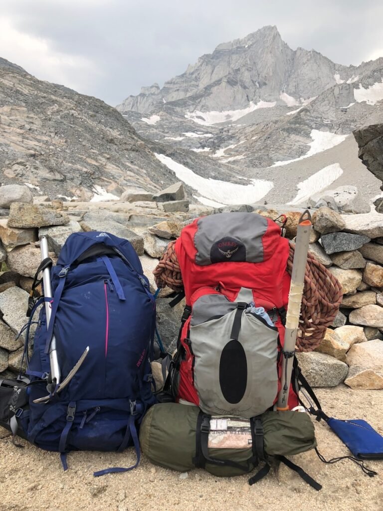 Two rugged backpack sits atop a woven Peruvian blanket, surrounded by essential wet-season gear - rain ponchos, hiking boots, and a waterproof camera. Misty mountains rise in the distance, their peaks shrouded in a soft, atmospheric haze. The lighting is natural and diffused, casting a warm, earthy glow across the scene. In the foreground, a map and compass lie ready, hinting at the adventure to come. This photo captures the careful preparation required for a Machu Picchu trek during the wet season, where weather and terrain demand thoughtful packing and planning.